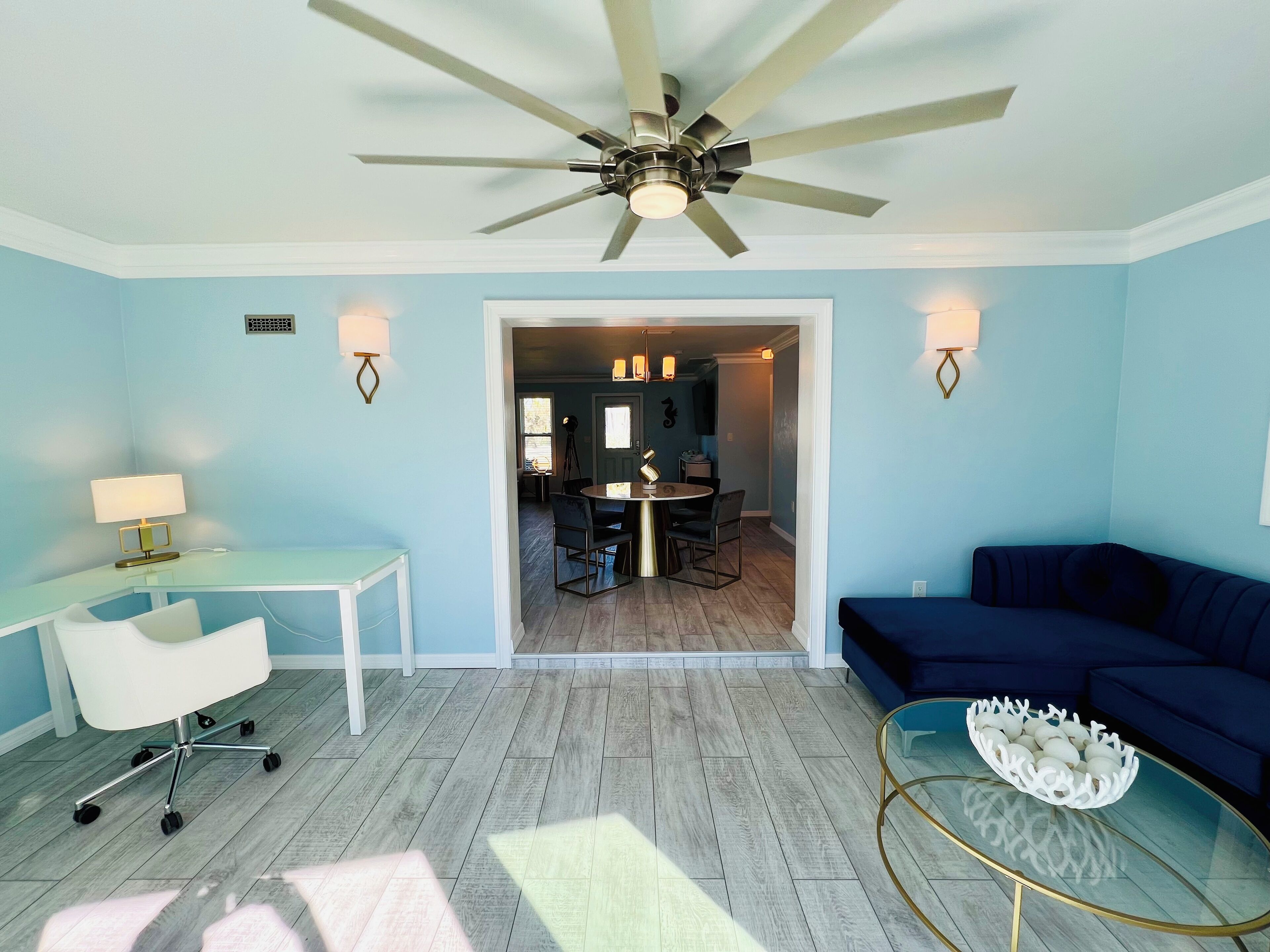 Une pièce avec un ventilateur de plafond, un bureau avec une lampe, un canapé bleu et une table basse en verre.