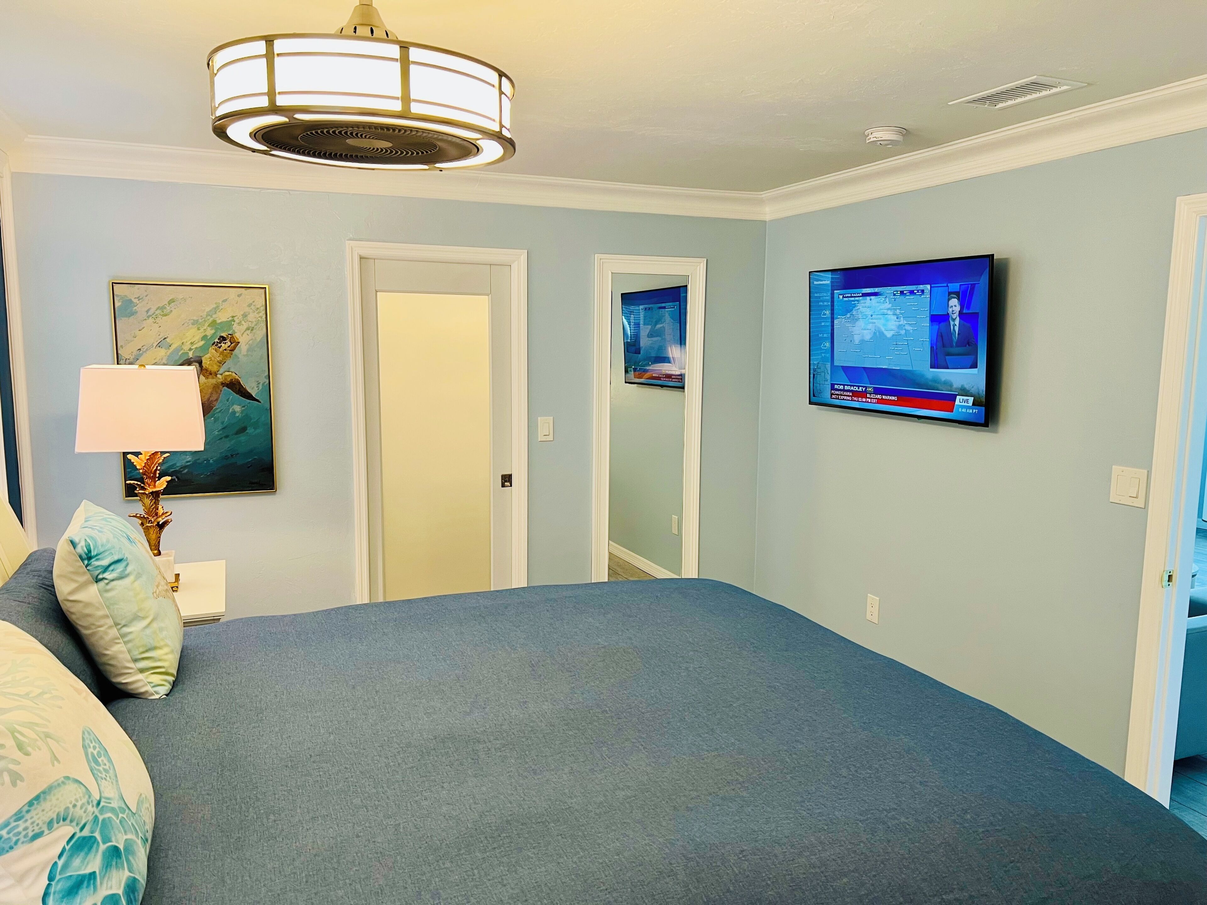 Un dormitorio con una cama grande, un televisor montado en la pared, un ventilador de techo y una puerta que da a otra habitación.