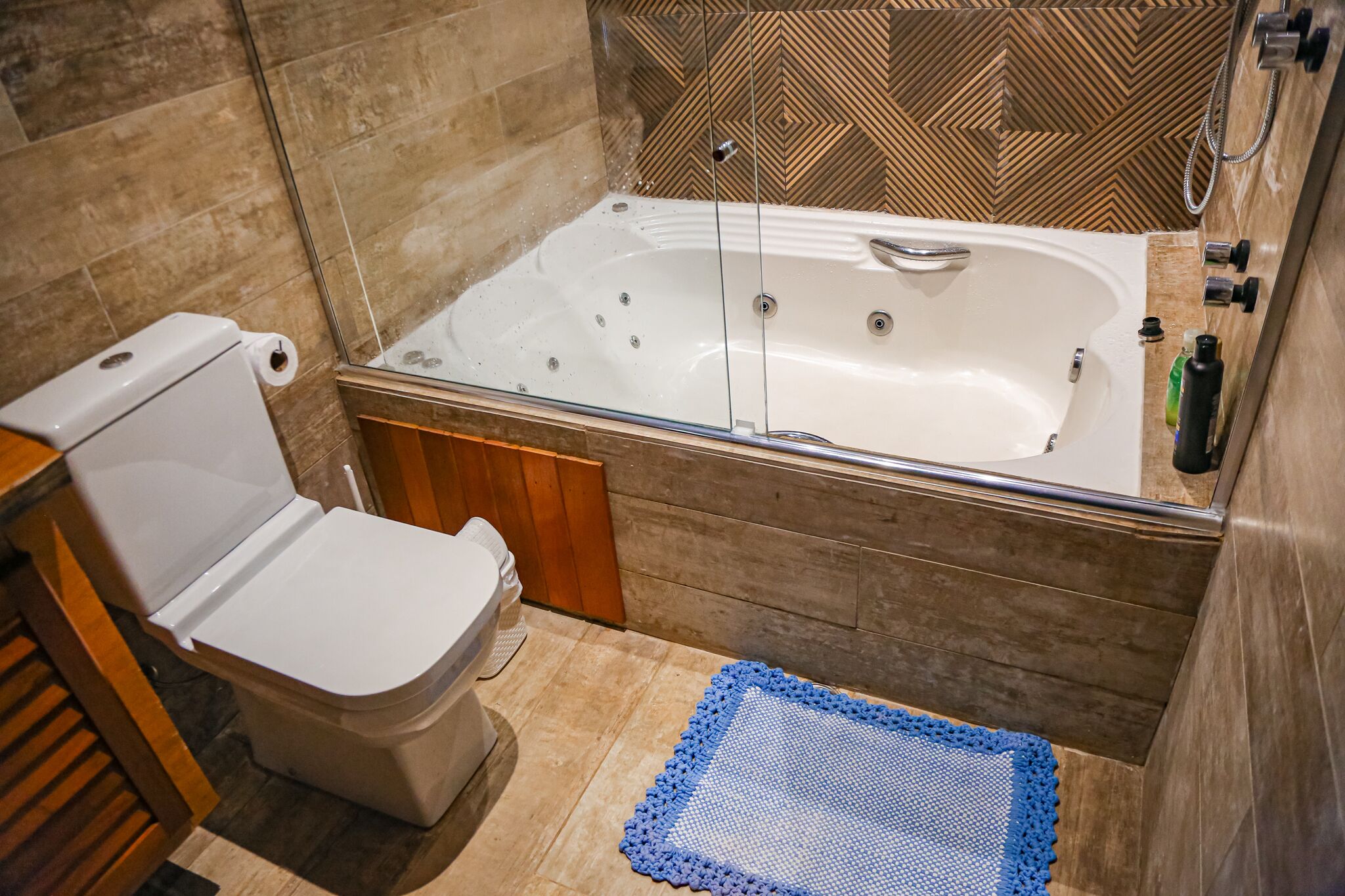 Un baño con inodoro, bañera con mampara de vidrio y un tapete azul con textura en el suelo.