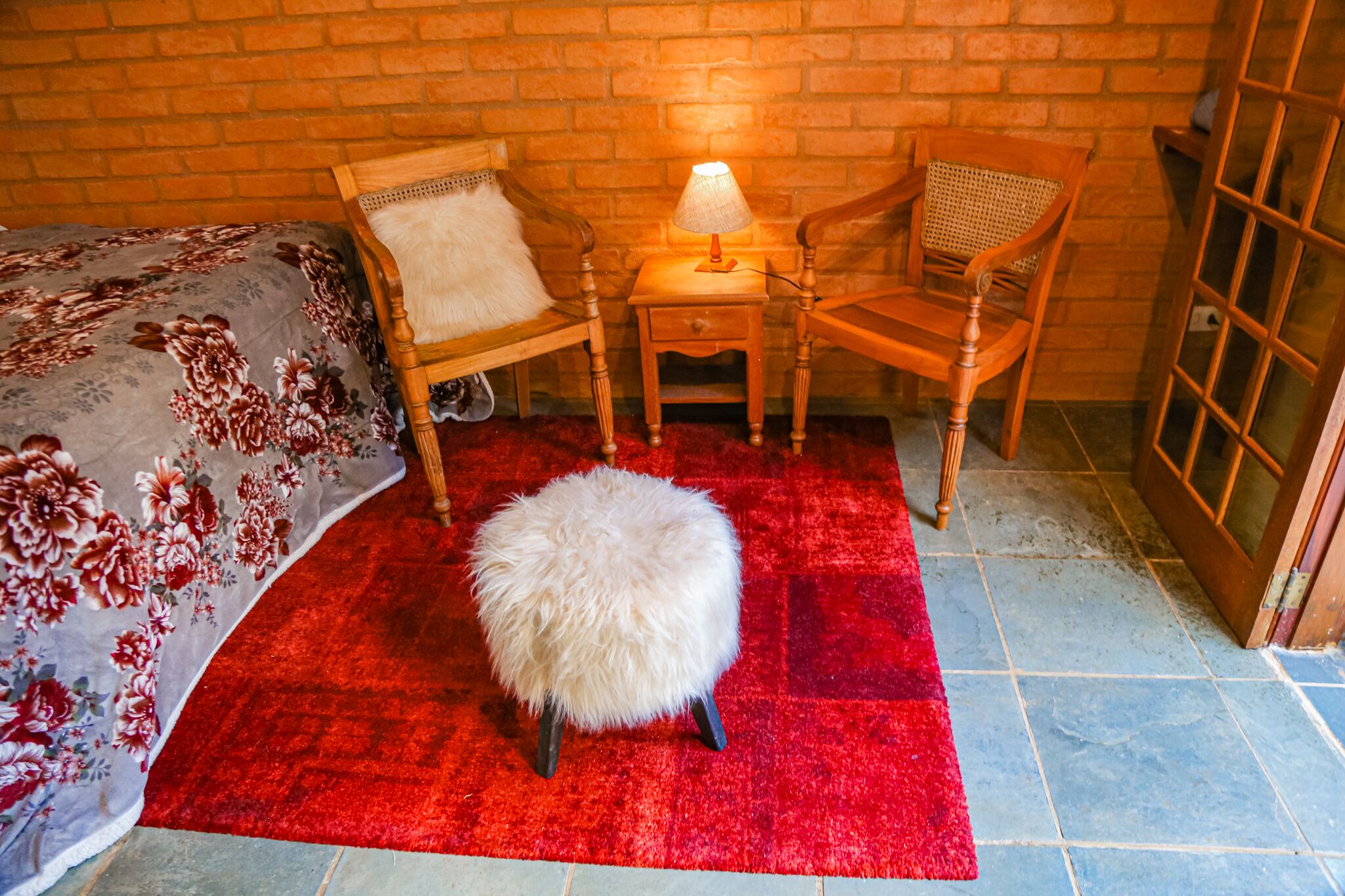 Une pièce avec un mur en briques, un lit recouvert d’une courtepointe à motifs floraux, deux chaises en bois, une petite table en bois avec une lampe, et un petit tabouret moelleux blanc posé sur un tapis rouge.