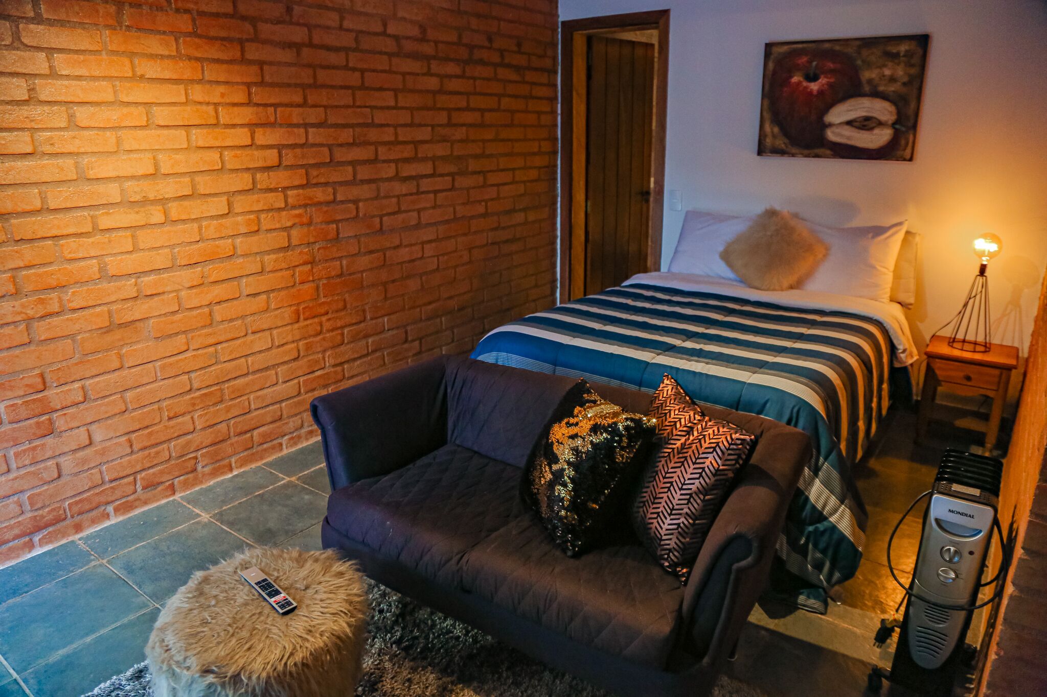 Ein Zimmer mit Backsteinwand, einem Bett mit gestreifter Bettwäsche, einer dunklen Couch mit gemusterten Kissen, einem kleinen runden Hocker und einem Nachttisch mit Lampe.