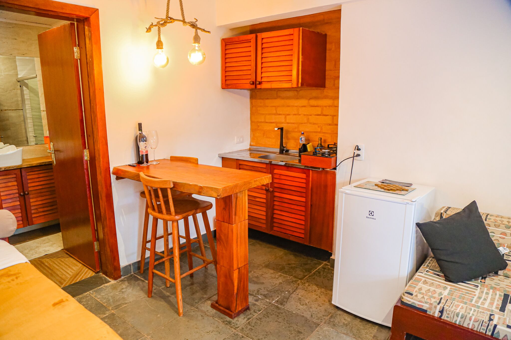 Een kleine keuken met houten kastjes, een kleine koelkast, een eettafel met stoelen en een bed met een kussen.