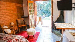 A room with a brick wall, a wooden door leading to a garden, a television mounted on the wall, and a bed with a floral bedspread. - Prime Estúdios (Campos do Jordão)