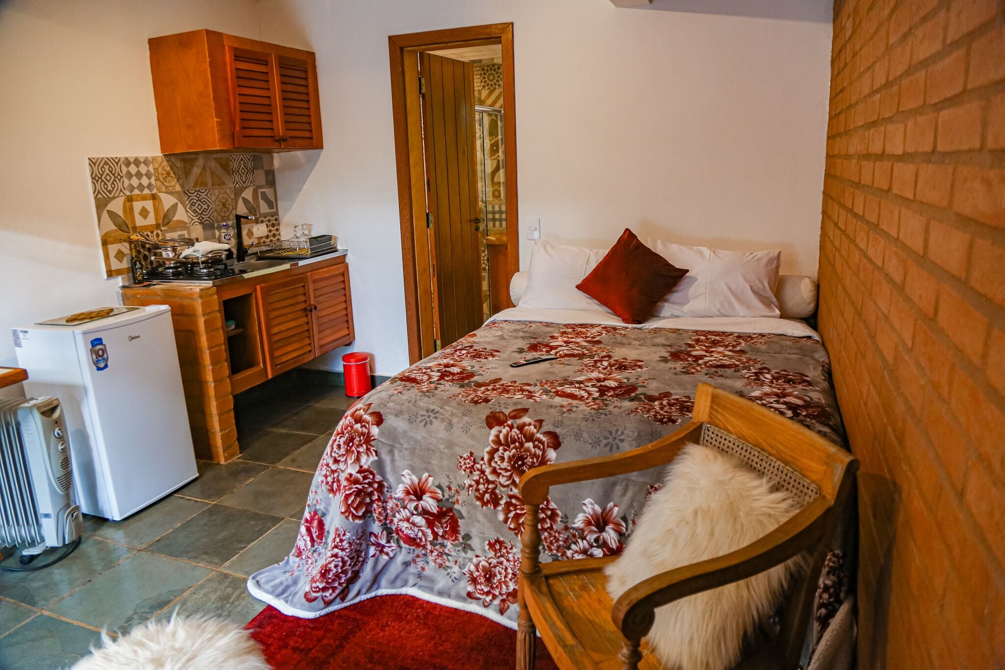 Een slaapkamer met een bed, een stoel, een klein tafeltje, een verwarming, een koelkast en een keukenhoek met kastjes en een fornuis.