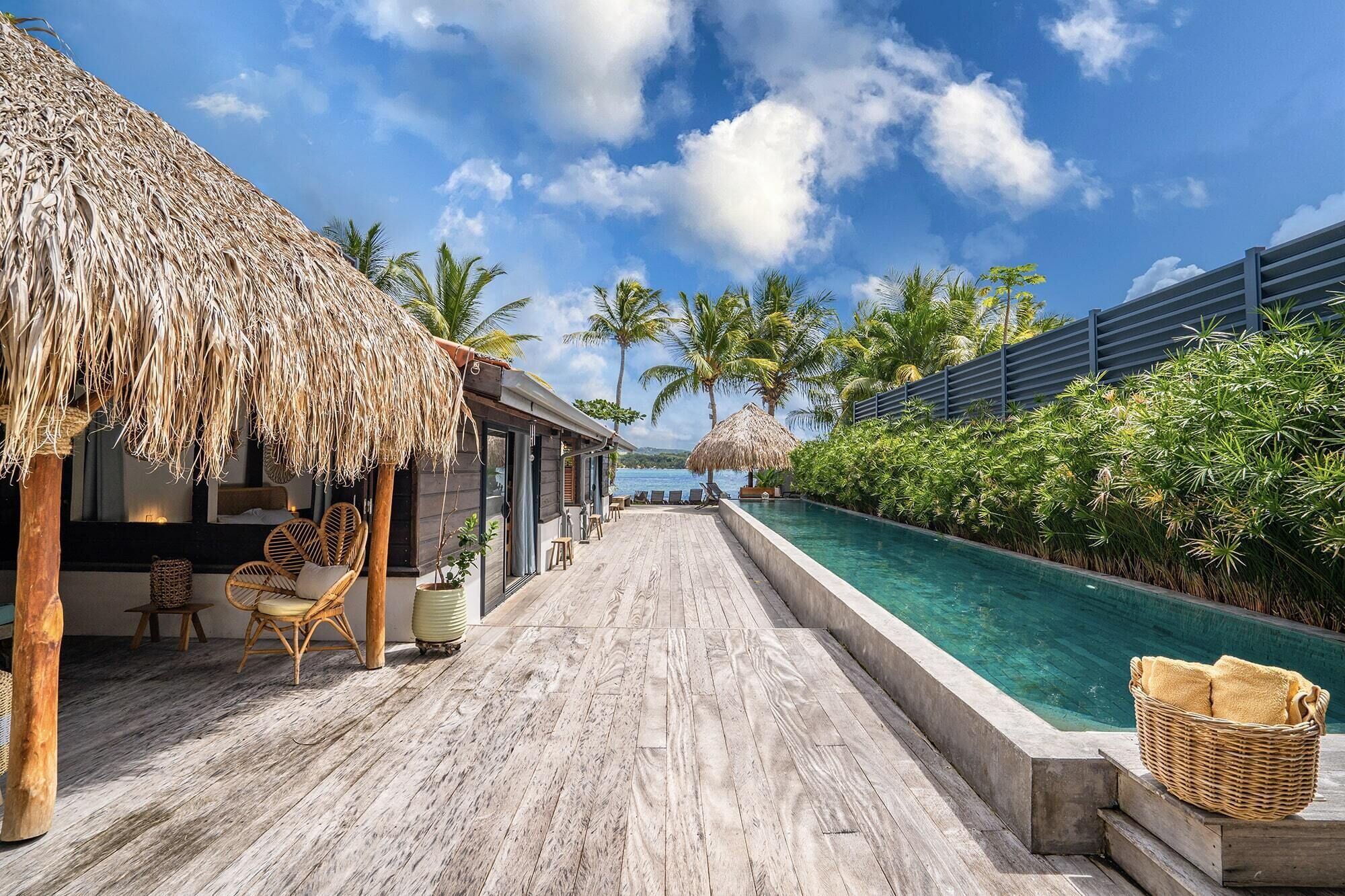 A wooden deck with a pool, thatched-roof structures, and palm trees.