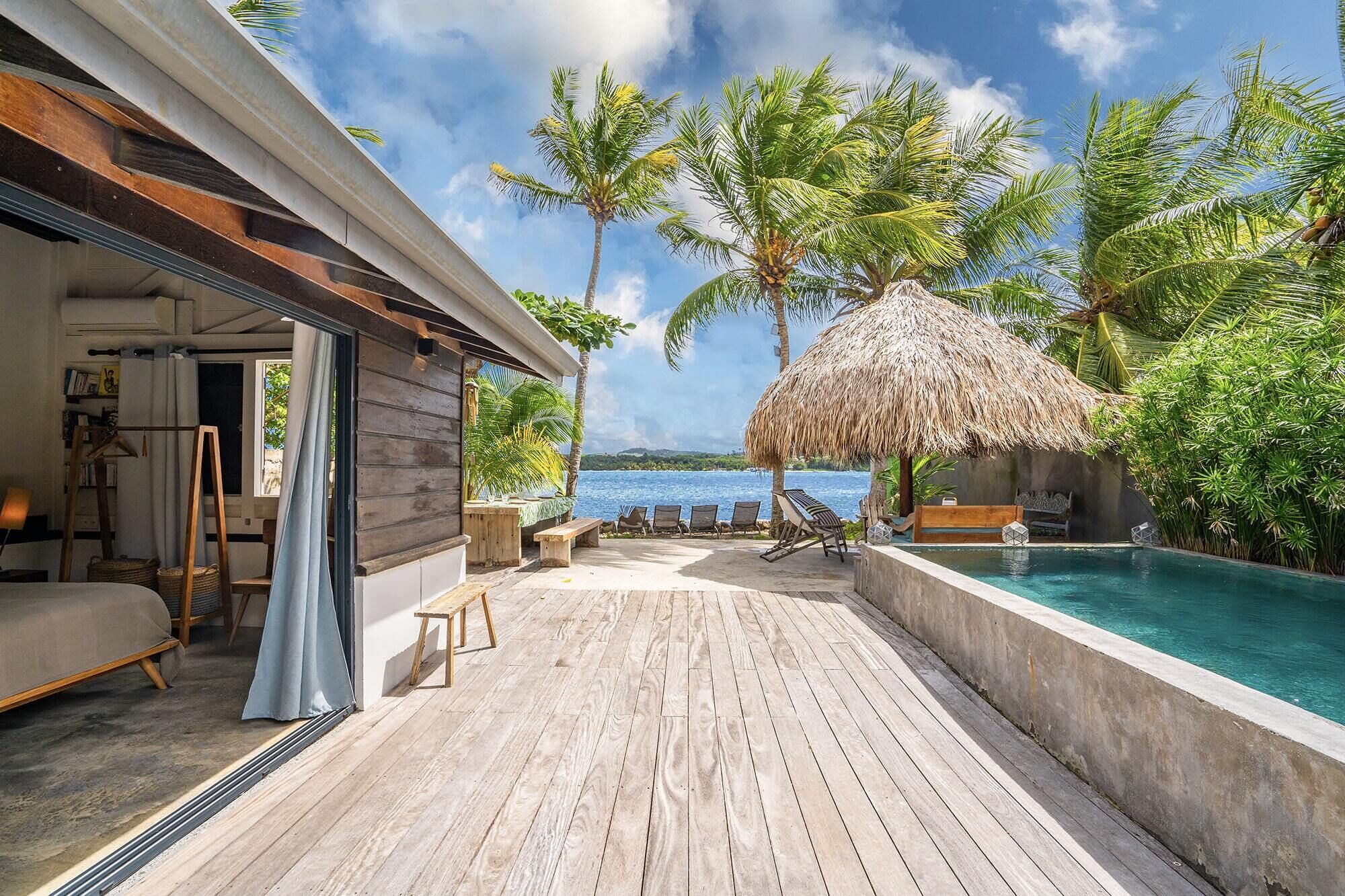 A wooden deck with a pool, a thatched umbrella, and palm trees.