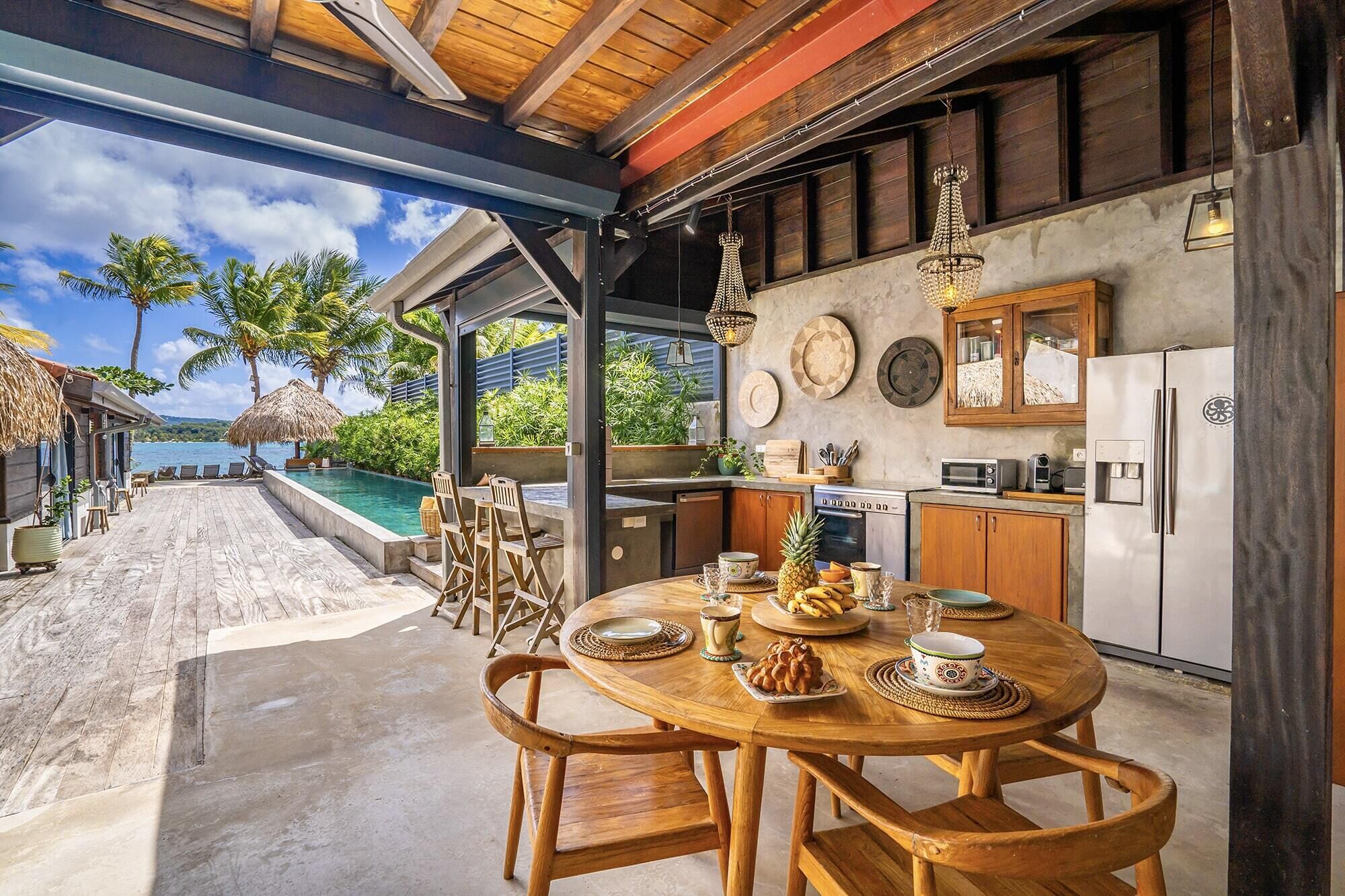 Ein Holzespeisesaal mit Stühlen, eine Küche mit Holzausstattung, ein Kühlschrank sowie Blick auf ein Schwimmbecken und Palmen.