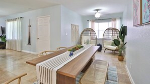 Un salón amplio con una mesa de comedor de madera, dos sillas de mimbre, un ventanal con cortinas y un ventilador de techo.