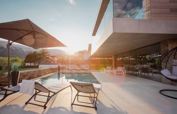 Un área de piscina moderna al aire libre con sillones de descanso y sombrillas, rodeada por un edificio contemporáneo con amplios ventanales.