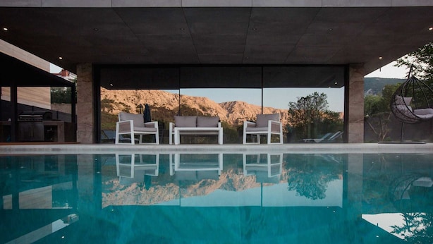 Un área de piscina moderna con paredes de vidrio transparente, sillones blancos y vistas a un paisaje montañoso.