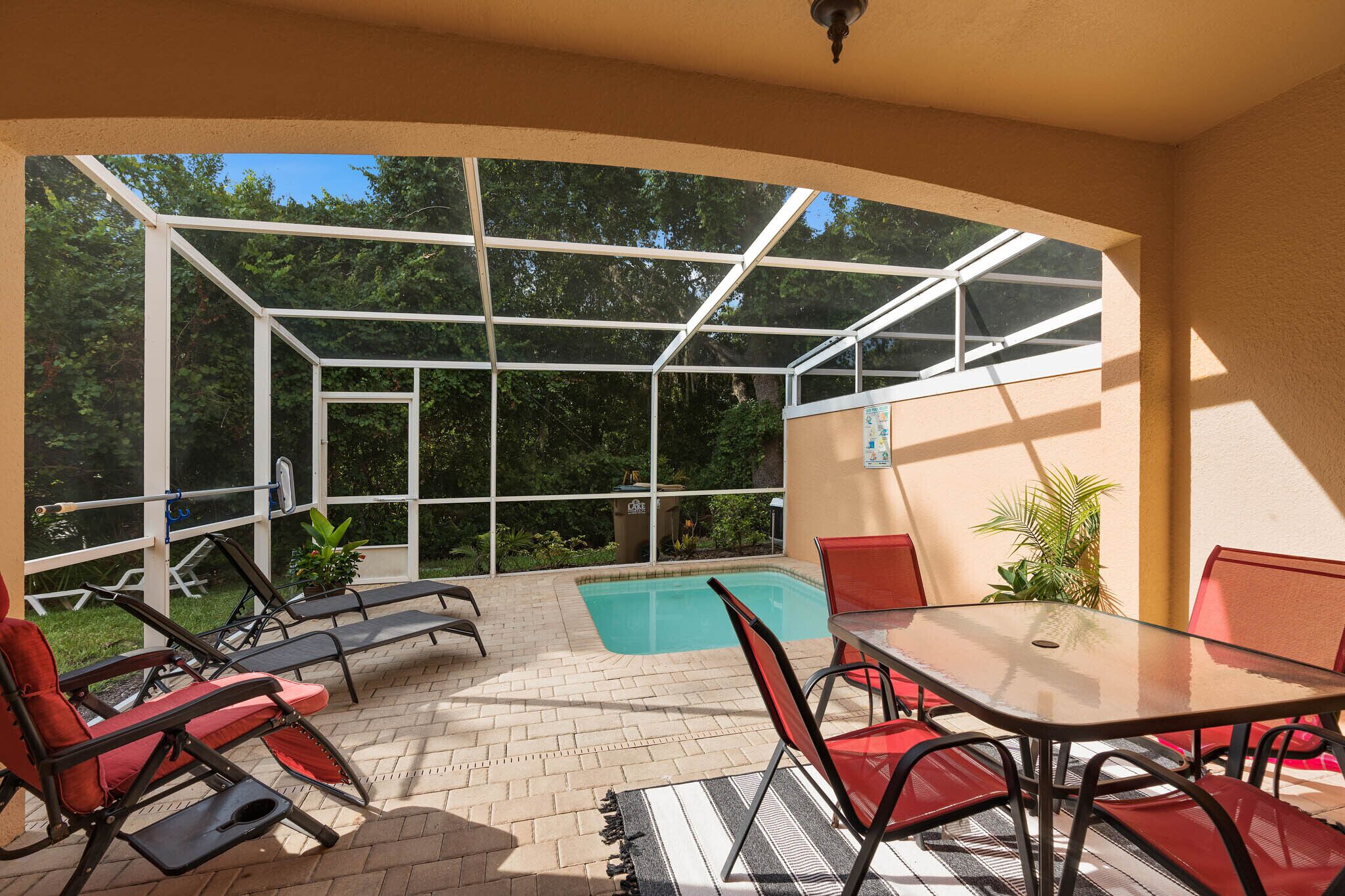 Área externa iluminada pelo sol com piscina envidraçada, cadeiras de lounge vermelhas e pretas, e uma mesa com cadeiras.