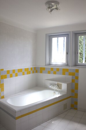 Une salle de bain avec une baignoire blanche, des carreaux jaunes et gris, une fenêtre et un luminaire au plafond.