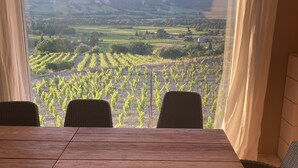 A dining area with a table and chairs, a view of vineyards through a large window, and a curtain on the right. - Villa provençale 6 pers, Piscine à débordement/Vue exceptionnelle/2 SDB, Ventoux (Villedieu)