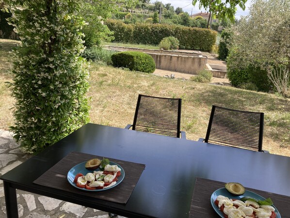 Une table à manger en plein air, avec deux assiettes de nourriture, entourée de chaises, dans un jardin offrant une vue sur un espace aménagé.