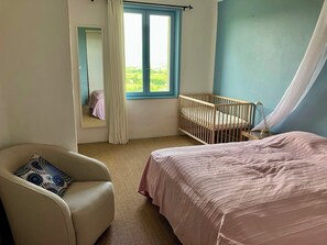 Une chambre avec un berceau, une chaise, un lit, une fenêtre avec des rideaux et une vue sur la verdure à l’extérieur.