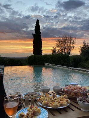 Un espace repas aménagé au bord de la piscine, offrant une vue sur le coucher du soleil, entouré de verdure et d’un grand arbre.