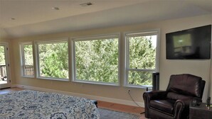 A bedroom with a large bed, a leather chair, a TV, and a view of trees through large windows. - Tree View - Murphys Vacation Rentals (Murphys)
