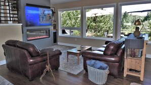 A living room with a flat-screen TV, leather sofas, a fireplace, and large windows overlooking a garden. - Tree View - Murphys Vacation Rentals (Murphys)