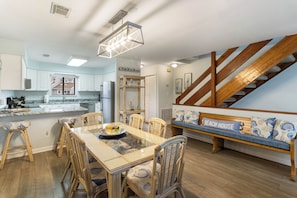 A modern kitchen with white cabinets, a dining table with chairs, a bench with a 'BEACH HOUSE' pillow, and a wooden staircase.