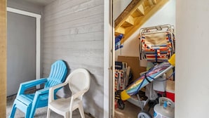 A small room with a wooden ceiling, a white door, and a window. There are two chairs, one blue and one white, and a bunk bed with striped bedding.