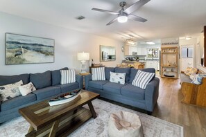 Un salon avec un canapé, une table basse et un pouf, un ventilateur de plafond, et une cuisine en arrière-plan.