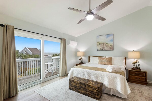 A bedroom with a bed, bedside lamps, a ceiling fan, a painting, and a view of a deck and house through a sliding door.