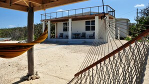 A hammock under a wooden structure, a building with a balcony, and a sandy beach in the background.