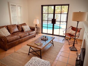 Ein Wohnzimmer mit braunem Ledersofa, einem Holztisch, einer Lampe und einem Sessel. Ein großes Fenster bietet Blick auf einen Pool und Palmen.
