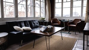 A modern living room with a black leather sofa, a wooden coffee table, and a dining area with two chairs. Large windows provide natural light and a view of the outside.
