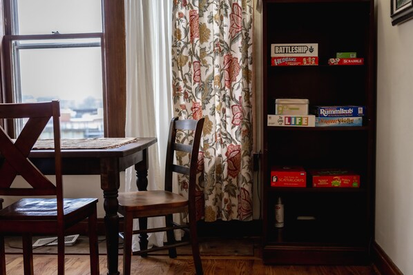 Une pièce avec une table et des chaises en bois, une bibliothèque remplie de jeux de société et une fenêtre avec des rideaux.
