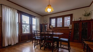 A dining room with a wooden table and chairs, large windows with white curtains, and a hanging light fixture.