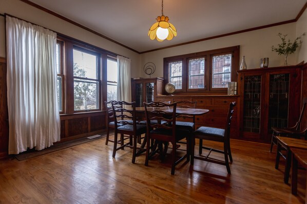 A dining room with a wooden table and chairs, large windows with white curtains, and a hanging light fixture.
