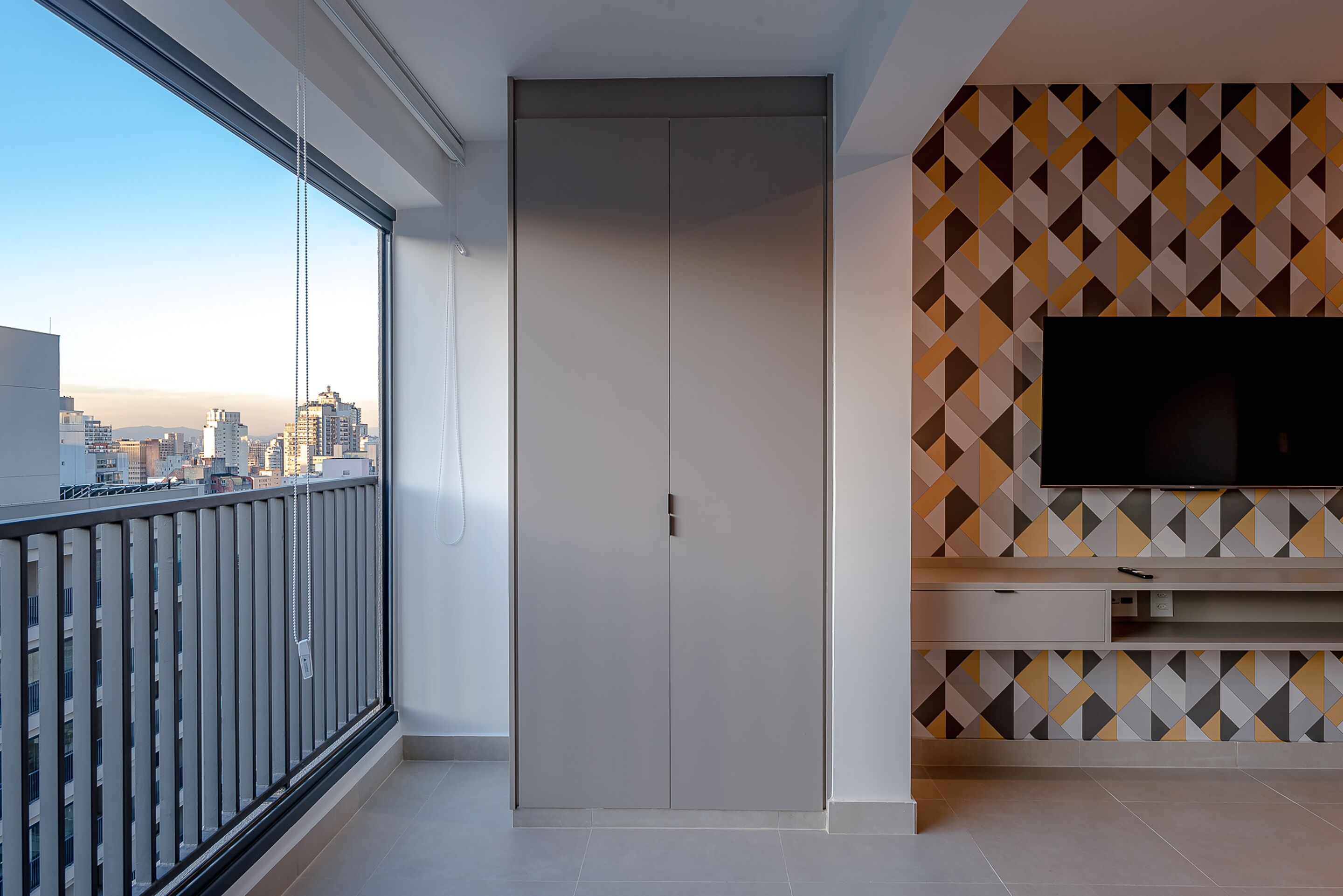 Eine moderne Wohnung mit großem Fenster, einem Kleiderschrank, einem Fernseher und einer Wand mit geometrischem Muster.