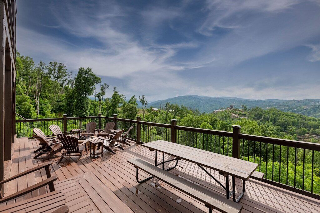 Una terraza de madera con sillas Adirondack y una mesa de picnic, con vistas a una cordillera boscosa.