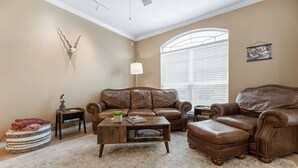 Un salon avec un canapé en cuir marron, un pouf assorti, une table basse en bois et une grande fenêtre avec des stores.