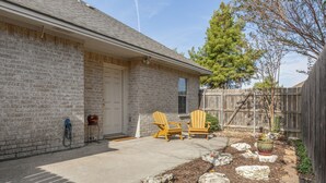 Une maison en briques avec une porte d’entrée, deux fauteuils Adirondack, un barbecue et un jardin avec du paillis et des rochers.