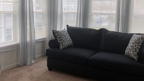Ein Wohnzimmer mit einem dunkelgrauen Sofa, gemusterten Kissen und leichten Vorhängen an großen Fenstern.