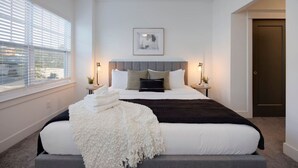 A modern bedroom with a large bed, two bedside tables, a framed picture on the wall, and a view through the window.