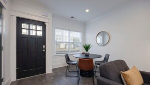 A modern living room with a dark wooden door, a round table with chairs, a grey sofa, and a round mirror on the wall.
