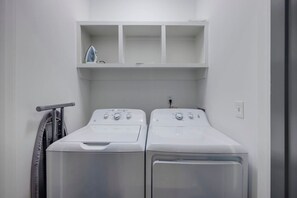 A laundry room with two front-loading washing machines, a drying rack, and a shelf above the machines.
