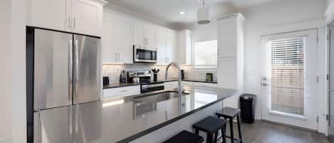 Una cocina moderna con armarios blancos, electrodomésticos de acero inoxidable, una isla grande con taburetes y un ventanal con persianas.