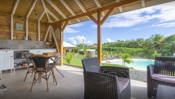 Ein überdachter Außenbereich mit Korbmöbeln, einer Küchenzeile und Blick auf Pool und Grünflächen.