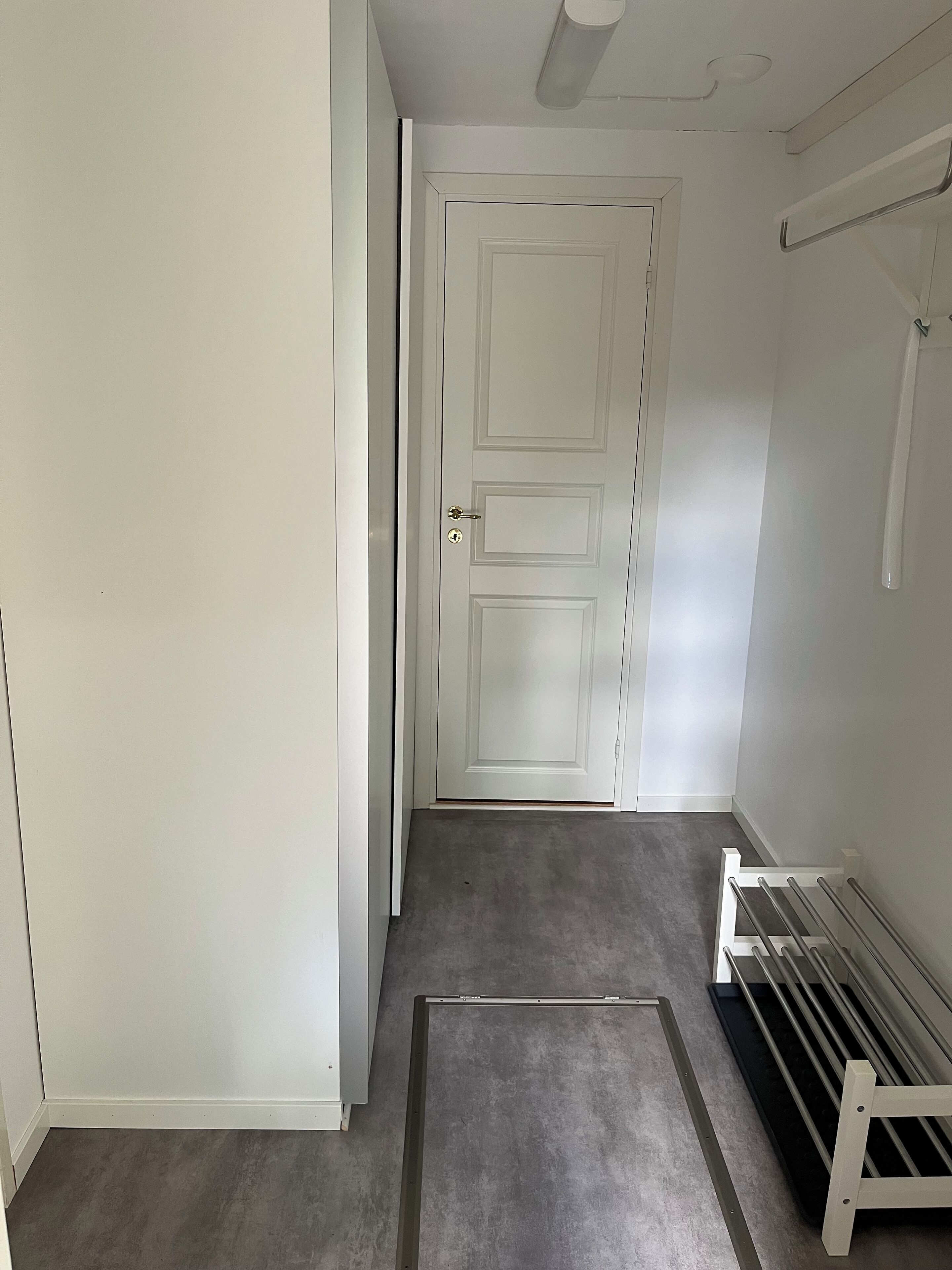 En hall med en vit dörr, ett grått golv och ett metallställ på väggen.