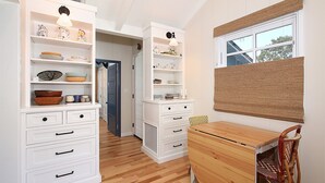 Una cocina con armarios blancos, suelo de madera y una ventana con persiana marrón.