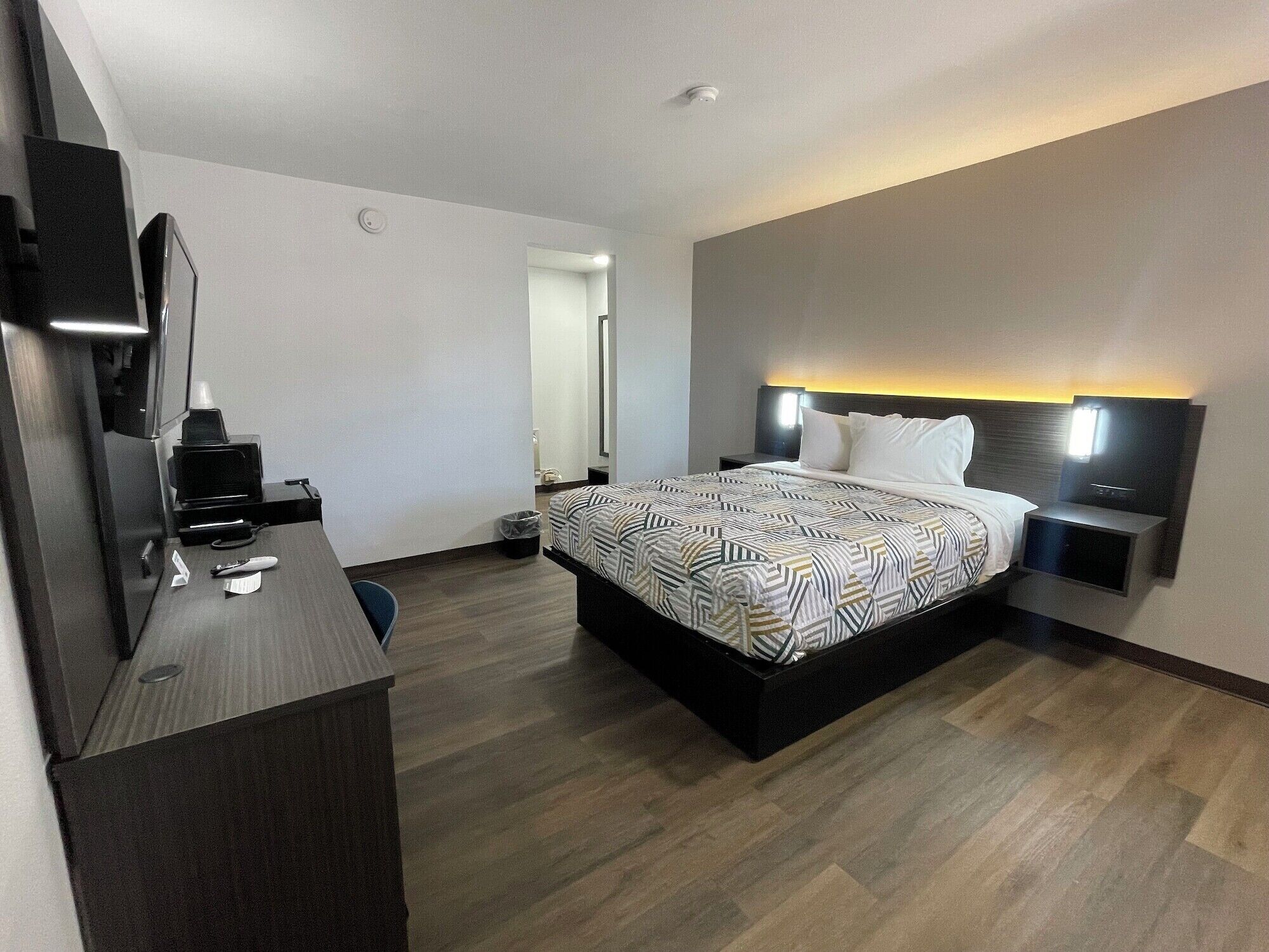A modern hotel room with a large bed, a desk with a chair, a flat-screen TV, and a bedside table with a lamp.