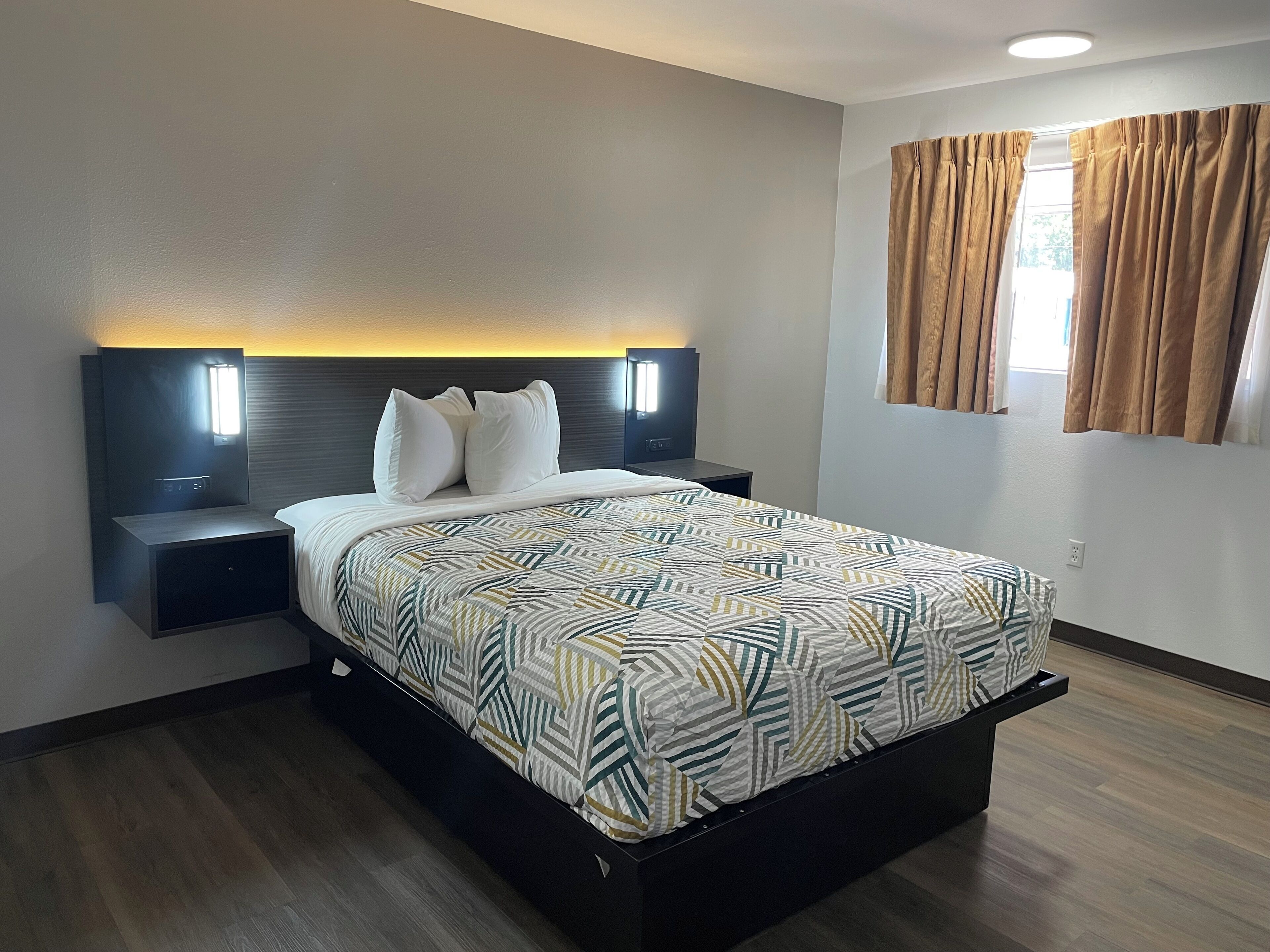 A modern hotel room with a large bed, two bedside tables with lamps, a headboard with built-in lighting, and a window with curtains.