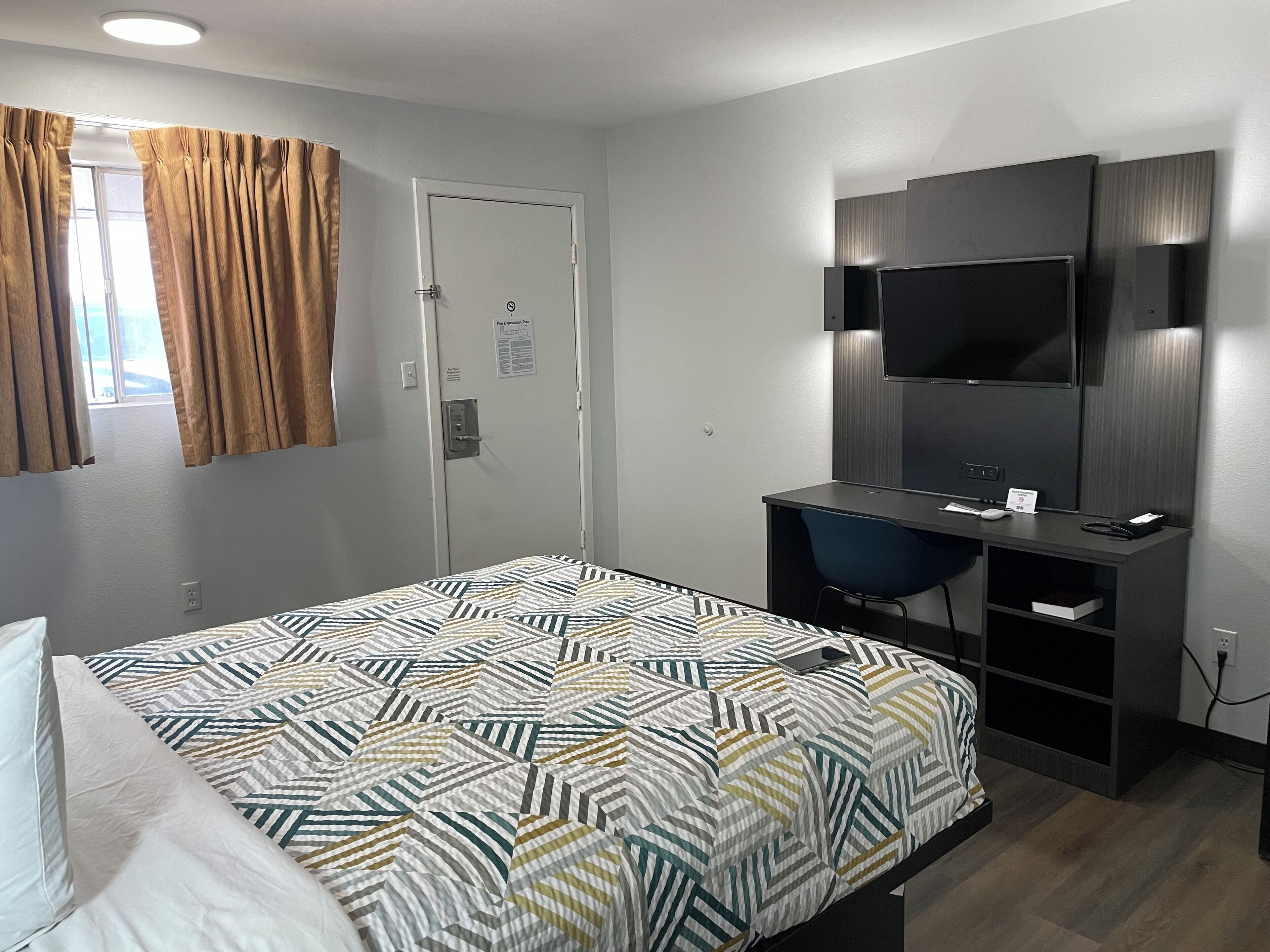 Une chambre d’hôtel équipée d’un lit, d’un bureau, d’une chaise, d’une télévision et d’une porte signalant la sortie de secours en cas d’incendie.