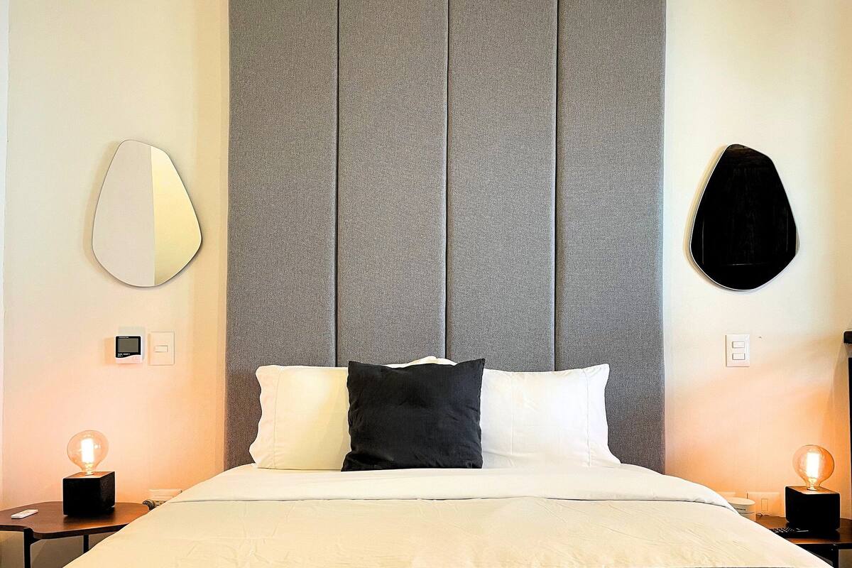 A modern hotel room with a large bed, a round mirror, and a unique textured headboard.