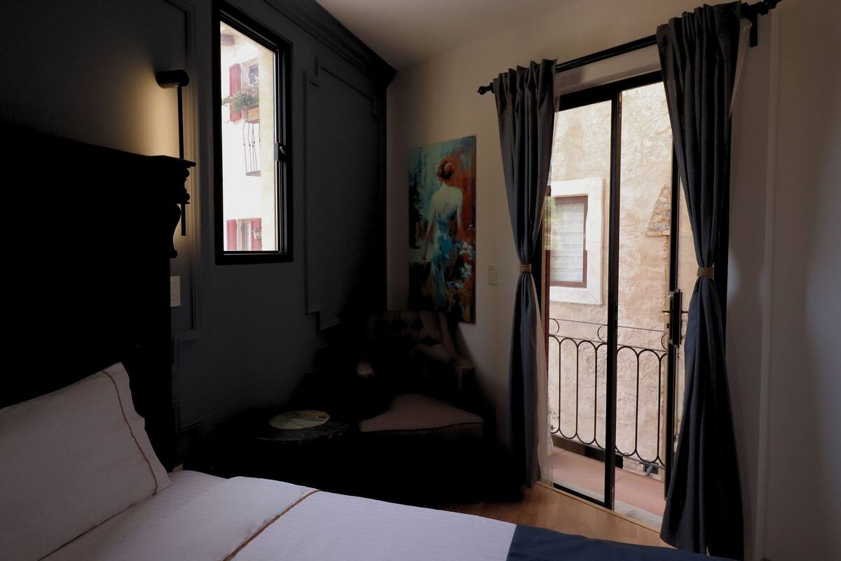 A bedroom with a bed, a chair, a painting, and a balcony with a view of buildings.