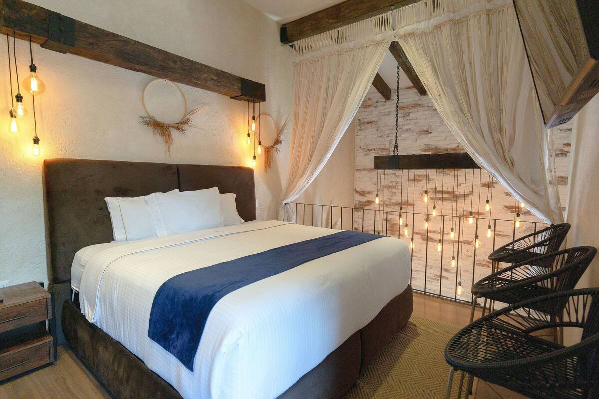 A bedroom with a large bed, a wooden headboard, a nightstand, and a chair. There are string lights on the wall and a curtain.